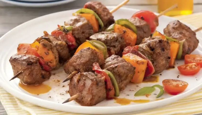 Brochettes de bœuf marinées, garnies de poivrons et tomates, sur une assiette blanche.