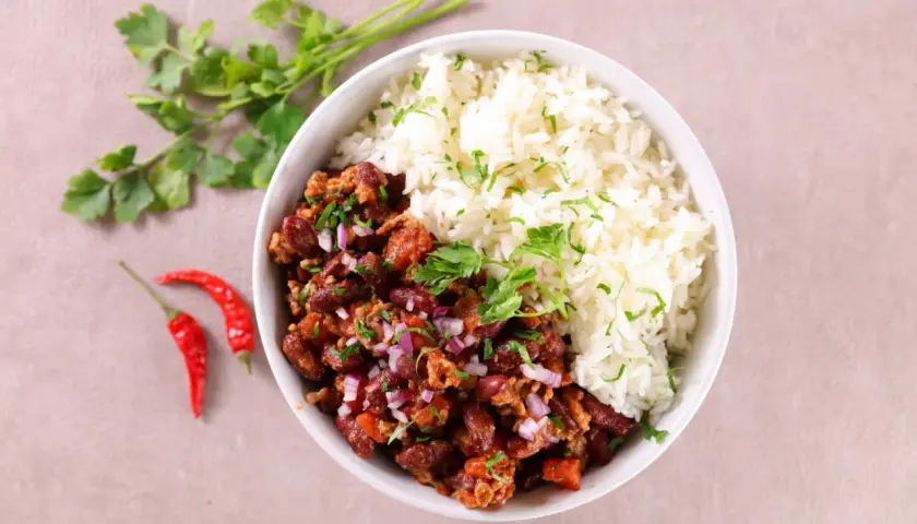 Un bol de chili con carne et riz, garni de persil frais et d'oignons rouges. Deux piments rouges sont posés à côté.
