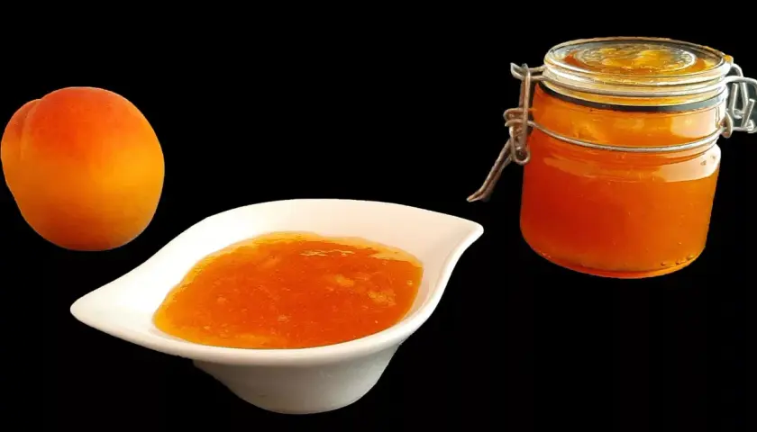 Un abricot frais à côté d'une coupelle de confiture abricot peu sucrée et d'un bocal de cette même confiture.
