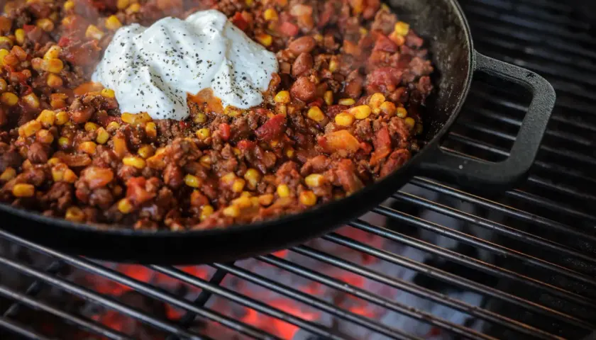 Chili con carne fumant dans une poêle en fonte sur un barbecue, garni d'une cuillère de crème fraîche poivrée.