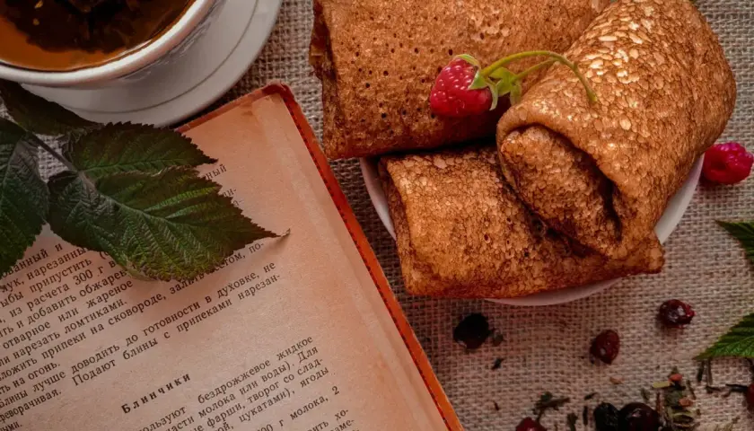 Crêpes roulées sur une assiette, accompagnées d'une tasse de thé et d'un livre de recette. Une framboise décore les crêpes. Parfait pour une recette crêpe.