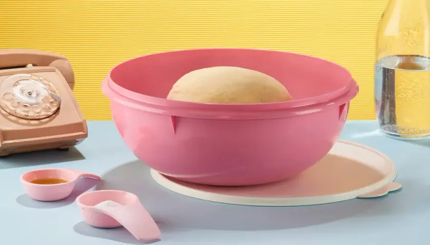 Pâte à pain dans un bol Tupperware rose, prêt pour une recette minute. À côté, un téléphone vintage, des cuillères doseuses et une bouteille d'eau.