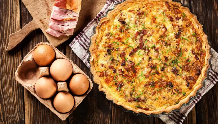 Une délicieuse quiche lorraine dorée, garnie de lardons et de ciboulette, accompagnée d'œufs frais et de lard fumé sur une planche en bois.