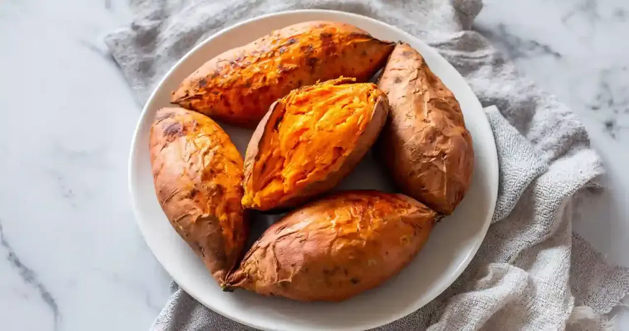 Cinq patates douces rôties à la perfection dans un air fryer, servies sur une assiette blanche. L'une est coupée en deux, révélant sa chair orange tendre.