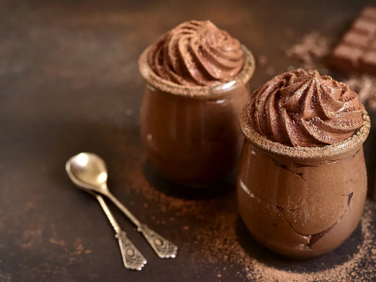 Deux verrines de recette mousse au chocolat au lait, saupoudrées de cacao, avec deux cuillères anciennes.