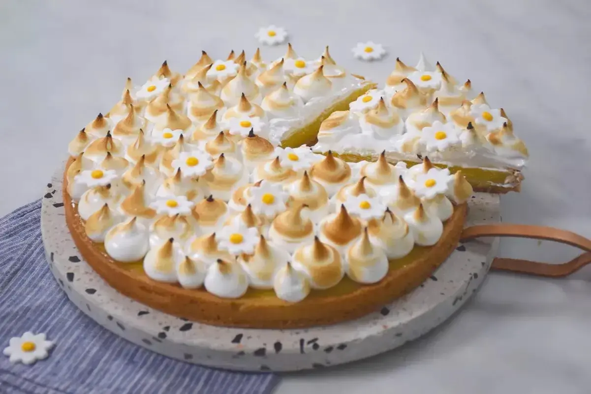 Une magnifique tarte aux citrons meringuée, décorée de petites fleurs blanches et jaunes, avec une part déjà coupée.