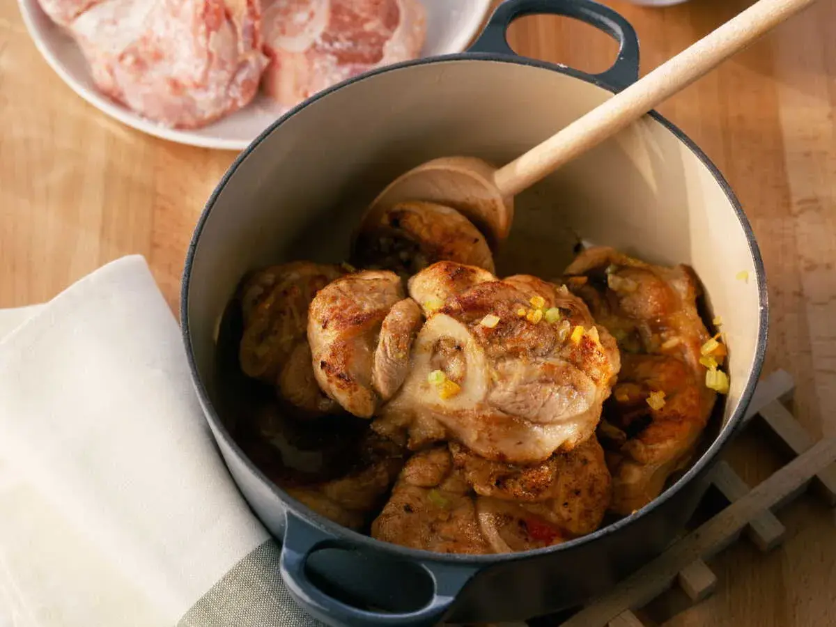 Jarret de veau mijoté dans une cocotte en fonte, garni d'herbes. Une cuillère en bois repose sur la viande.
