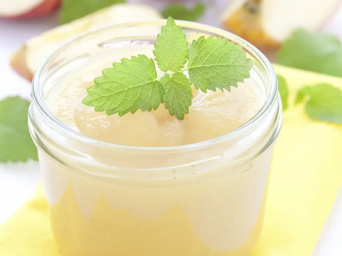 Un pot de compote pomme banane, garni d'une feuille de menthe fraîche. Des morceaux de pommes flous en arrière-plan.