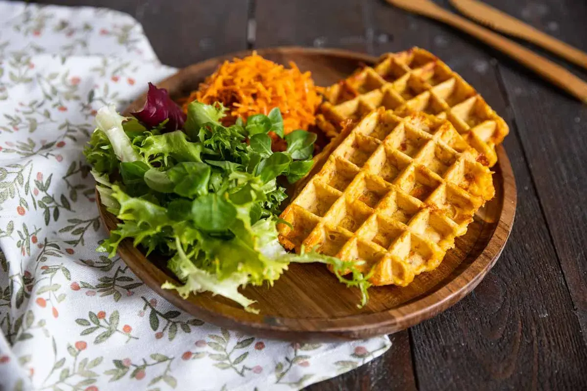 Gaufre patate douce dorée, servie avec une salade verte fraîche et une julienne de carottes râpées sur une assiette en bois.