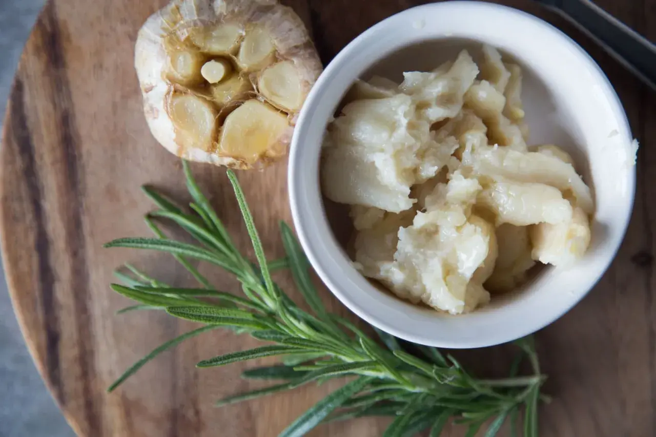 Ail confit au four, doux et crémeux, servi dans un bol blanc, à côté d'une tête d'ail rôtie et d'une branche de romarin frais.