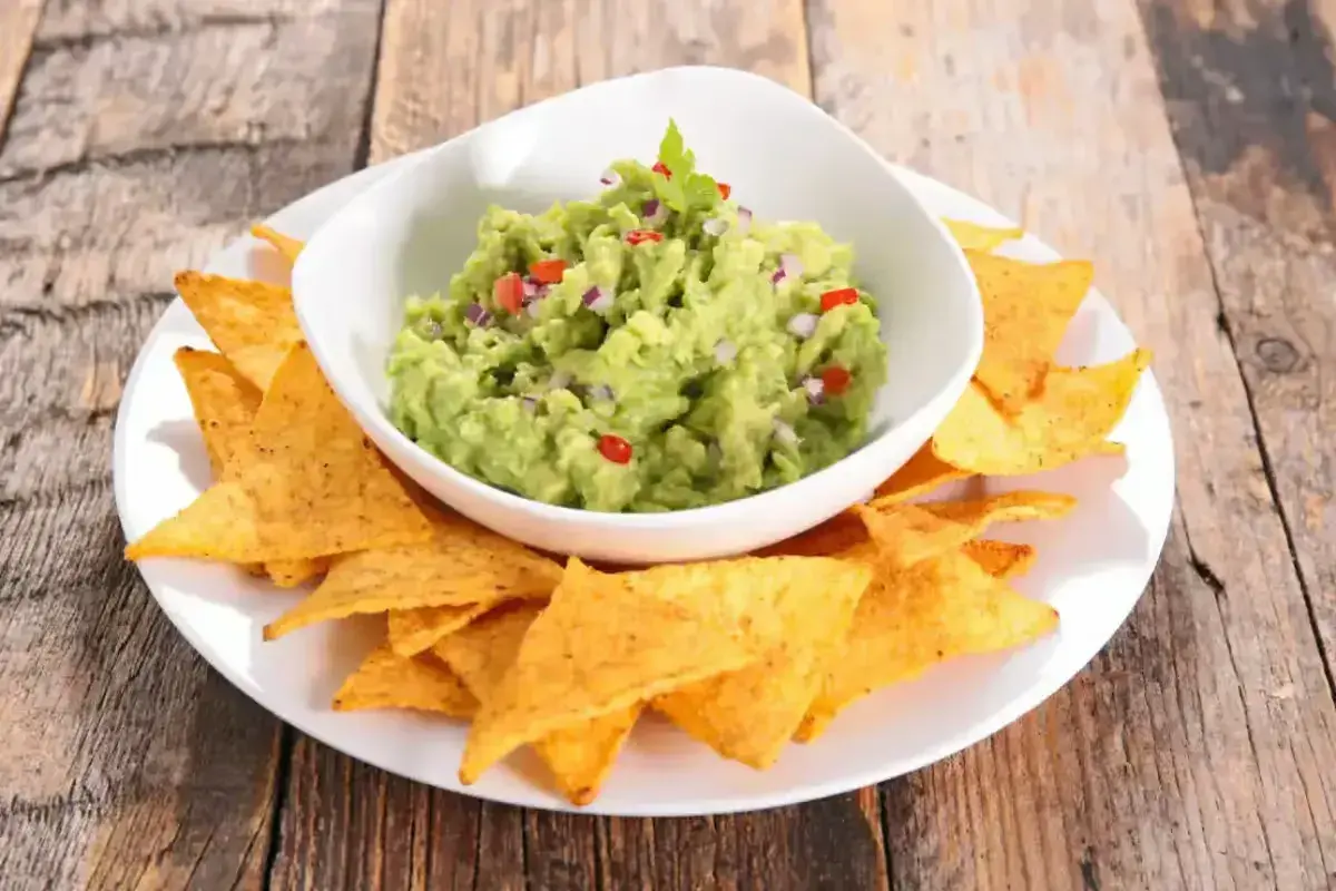 Un bol de guacamole traditionnel, garni de coriandre, est servi avec des chips tortilla sur une assiette blanche.