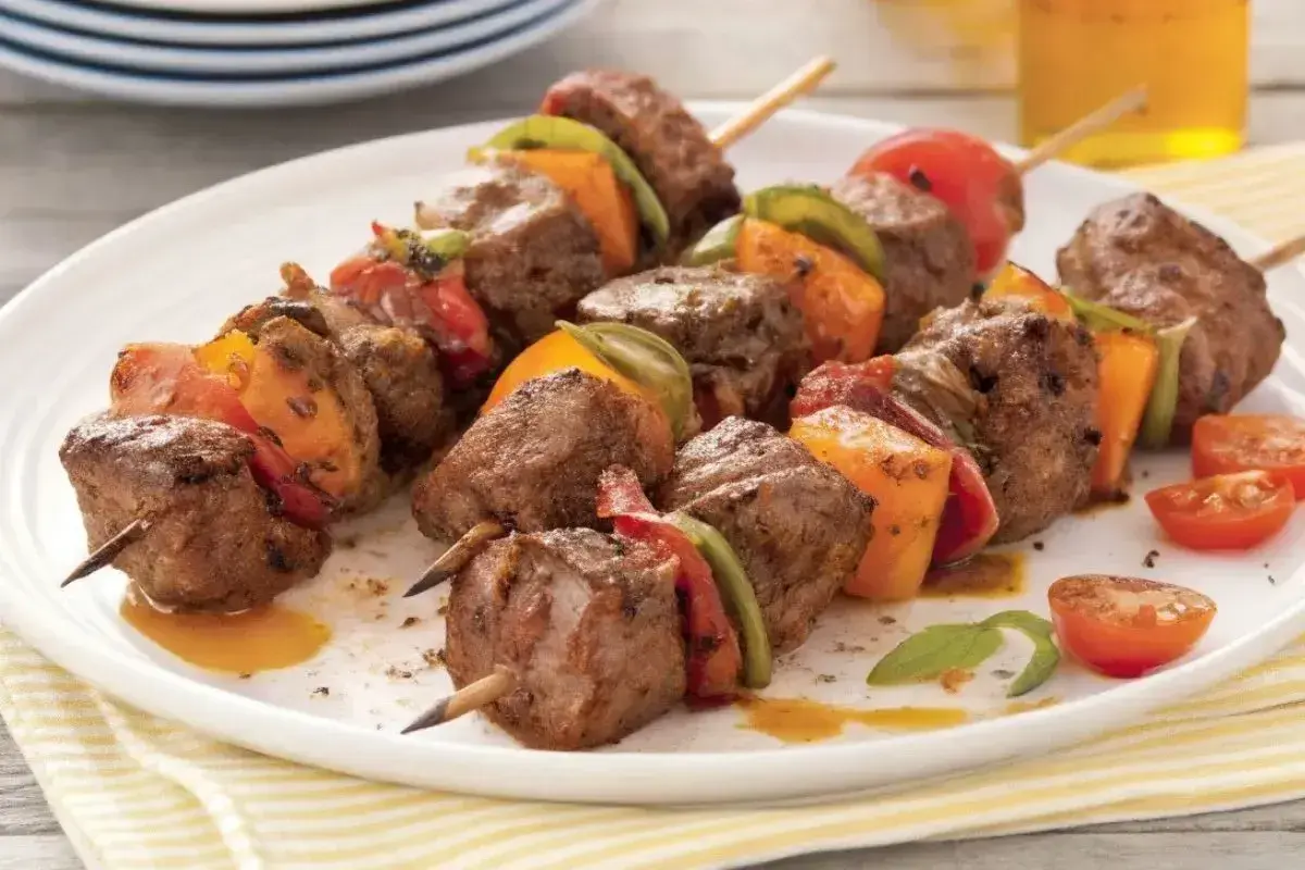 Brochettes de bœuf marinées, garnies de poivrons et tomates, sur une assiette blanche.
