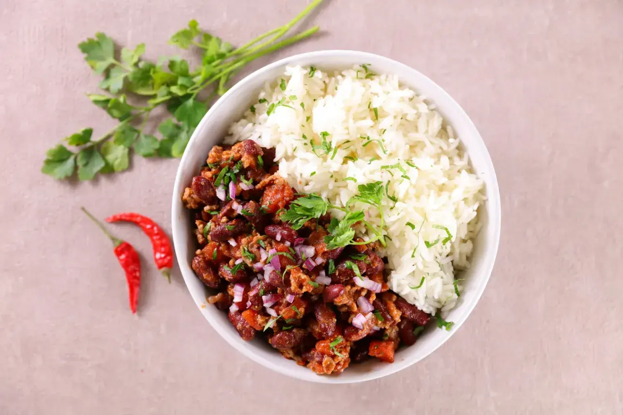 Un bol de chili con carne et riz, garni de persil frais et d'oignons rouges. Deux piments rouges sont posés à côté.