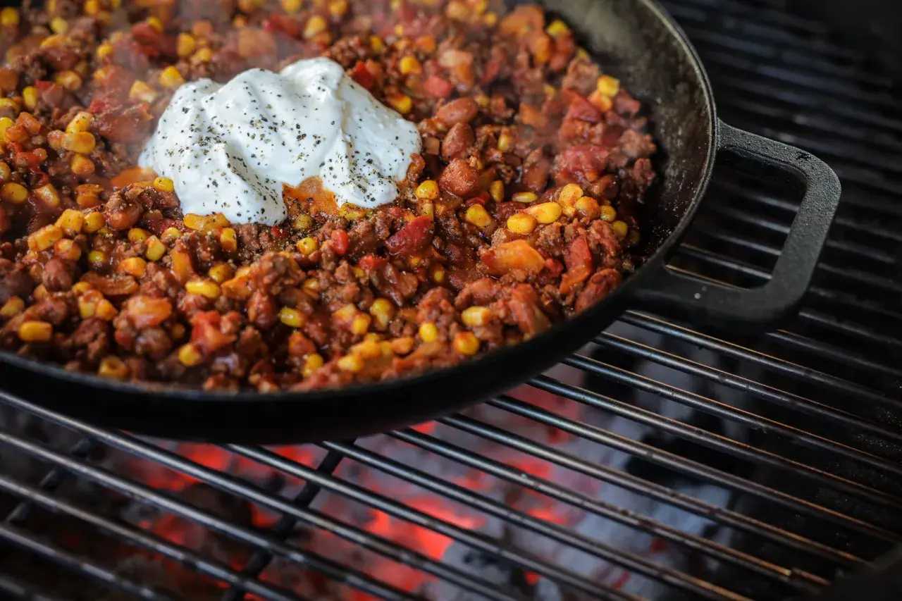 Chili con carne fumant dans une poêle en fonte sur un barbecue, garni d'une cuillère de crème fraîche poivrée.