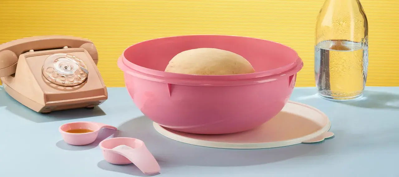 Pâte à pain dans un bol Tupperware rose, prêt pour une recette minute. À côté, un téléphone vintage, des cuillères doseuses et une bouteille d'eau.