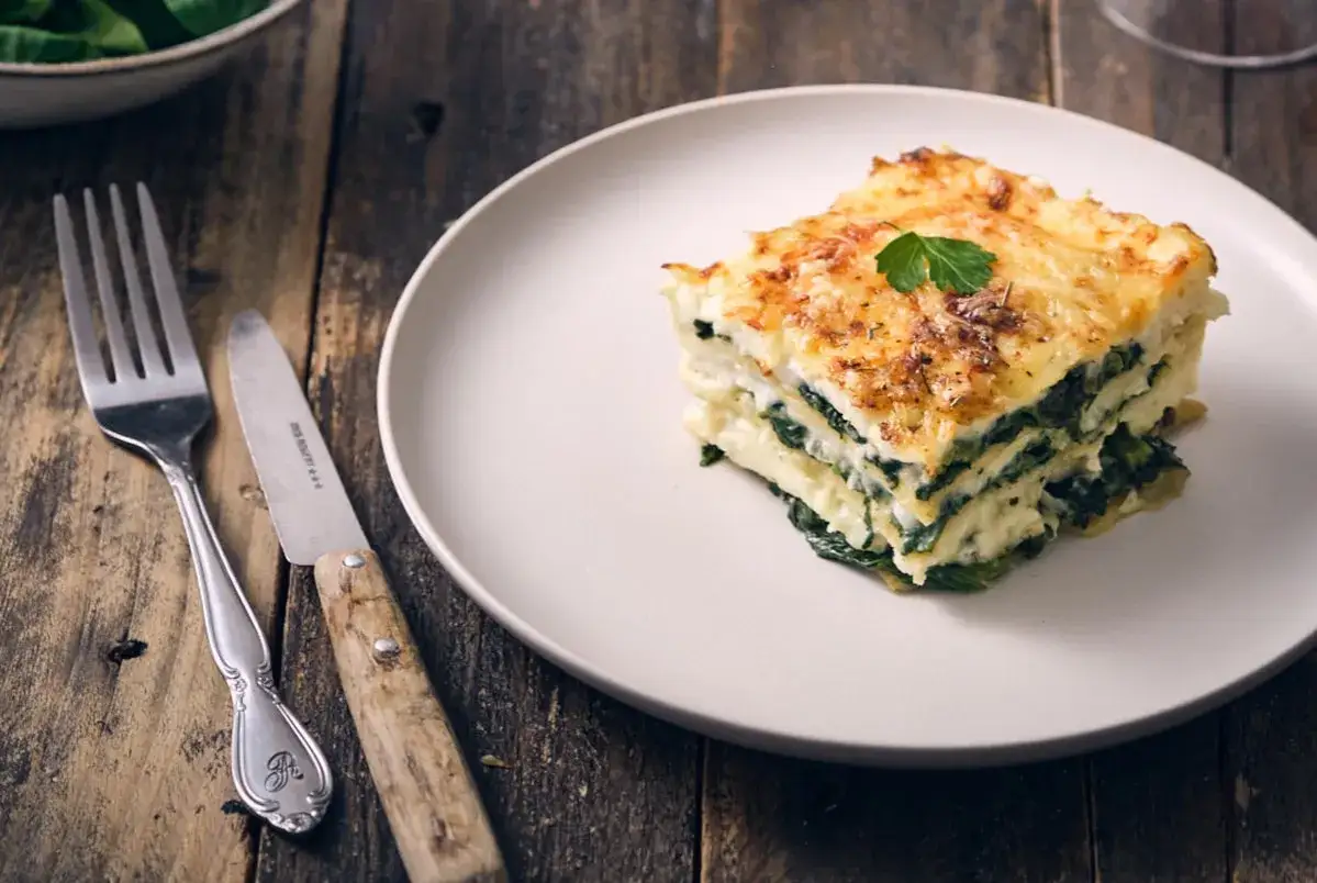 Une part de lasagne &eacute;pinard ch&egrave;vre, dor&eacute;e au four, sur une assiette blanche, accompagn&eacute;e d'une fourchette et d'un couteau sur fond de bois.