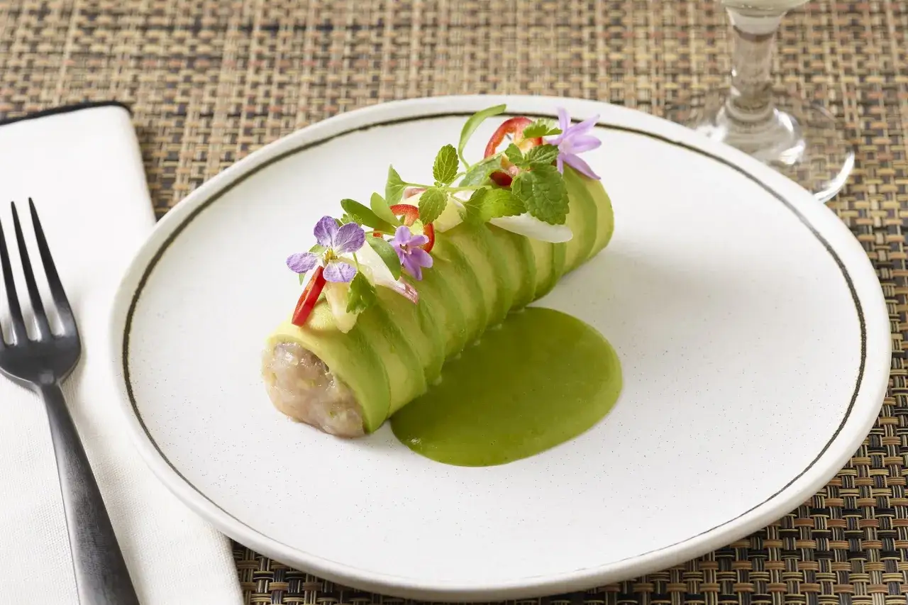 Un rouleau de tartare de thon frais, enveloppé d'avocat tranché, garni de fleurs comestibles et servi avec une sauce verte vibrante.