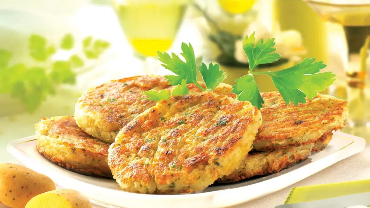 Pile de galettes de pomme de terre dorées, garnies de persil frais, sur une assiette blanche. Des pommes de terre crues sont visibles au premier plan.