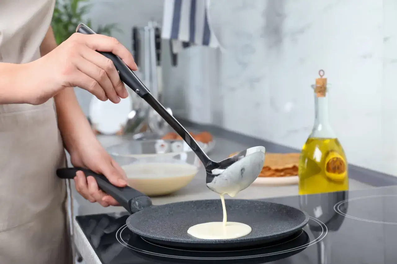Une louche verse de la pâte à crêpe dans une poêle chaude. Une bouteille d'huile et une pile de crêpes sont visibles en arrière-plan.