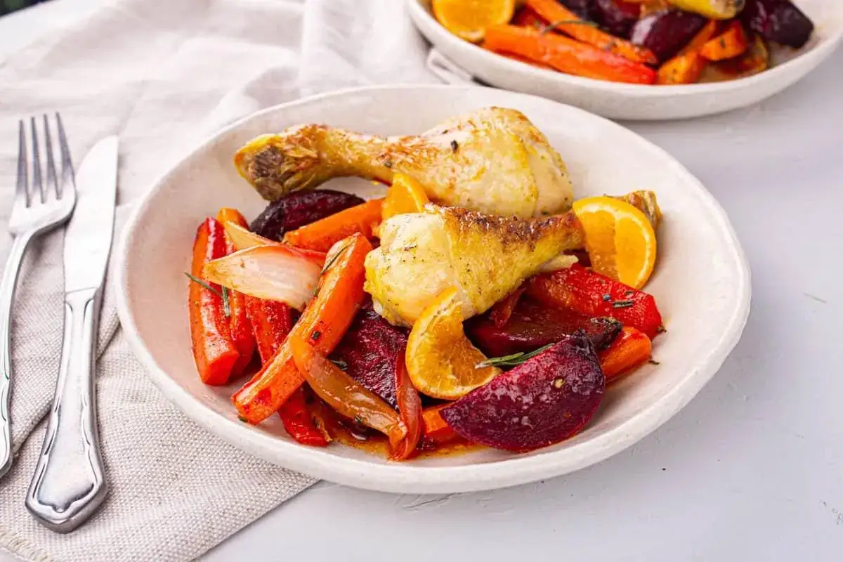 Un d&eacute;licieux pilon de poulet r&ocirc;ti avec des carottes, des betteraves, des oignons et des quartiers d'orange, le tout servi dans une assiette blanche.