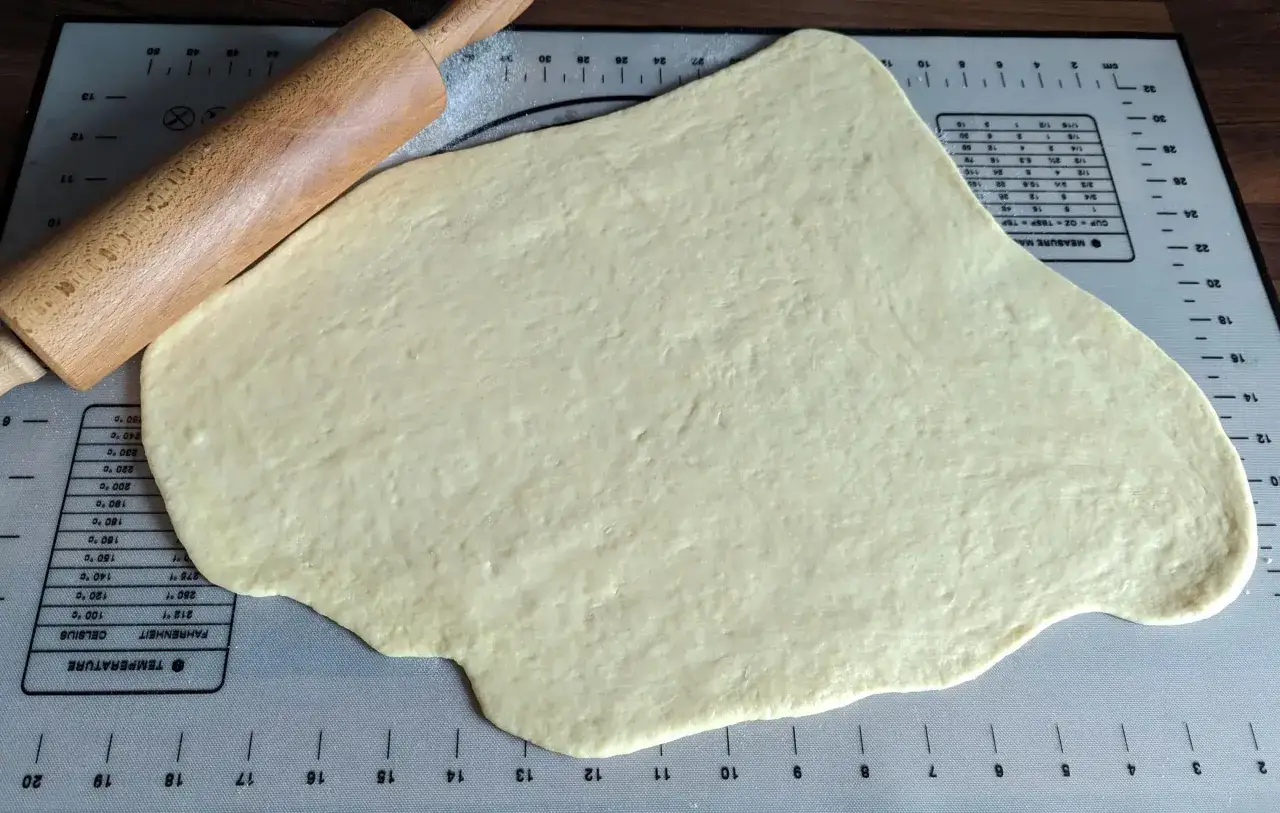 Pâte à beignet étalée sur un tapis de mesure, avec un rouleau à pâtisserie en bois.