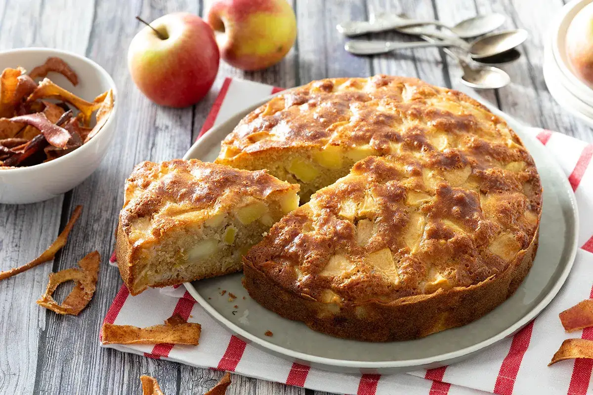 Un d&eacute;licieux g&acirc;teau aux pommes, dor&eacute; et moelleux, avec des morceaux de fruits visibles. Des pommes fra&icirc;ches et des chips de pommes compl&egrave;tent la sc&egrave;ne.
