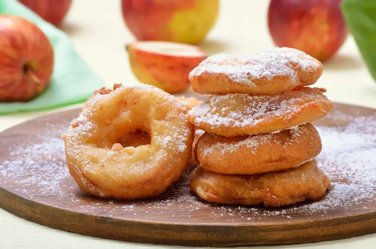 D&eacute;licieux beignets aux pommes &agrave; lancienne, saupoudr&eacute;s de sucre glace. Des pommes fra&icirc;ches en arri&egrave;re-plan rappellent la douceur de cette recette.