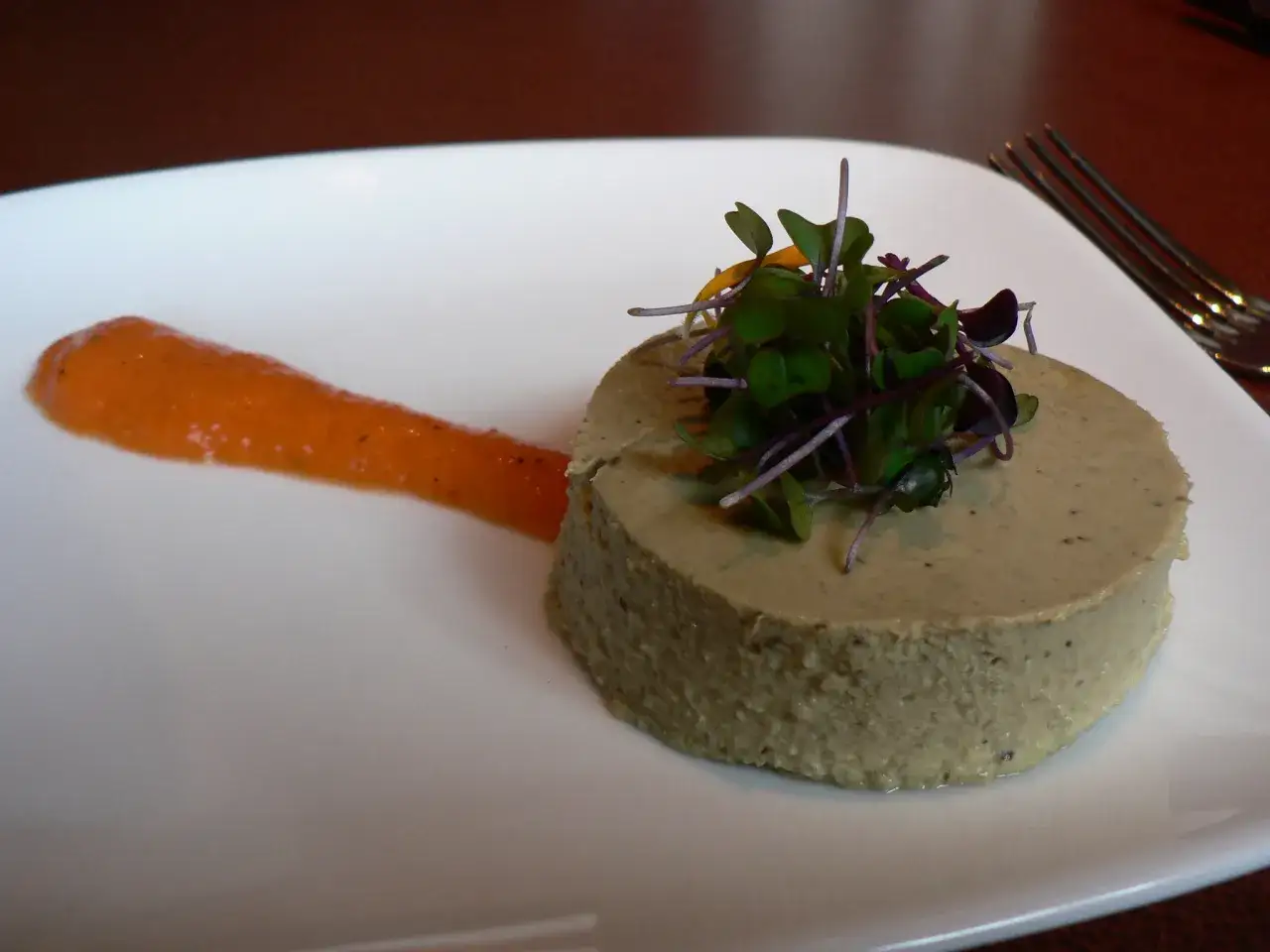 Un délicieux flan de légumes, garni de jeunes pousses, servi avec une sauce orangée sur une assiette blanche.