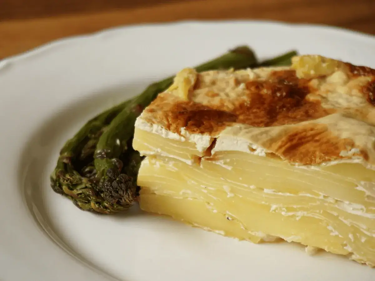 Tranche de gratin dauphinois dorée et crémeuse, accompagnée d'asperges vertes sur une assiette blanche.