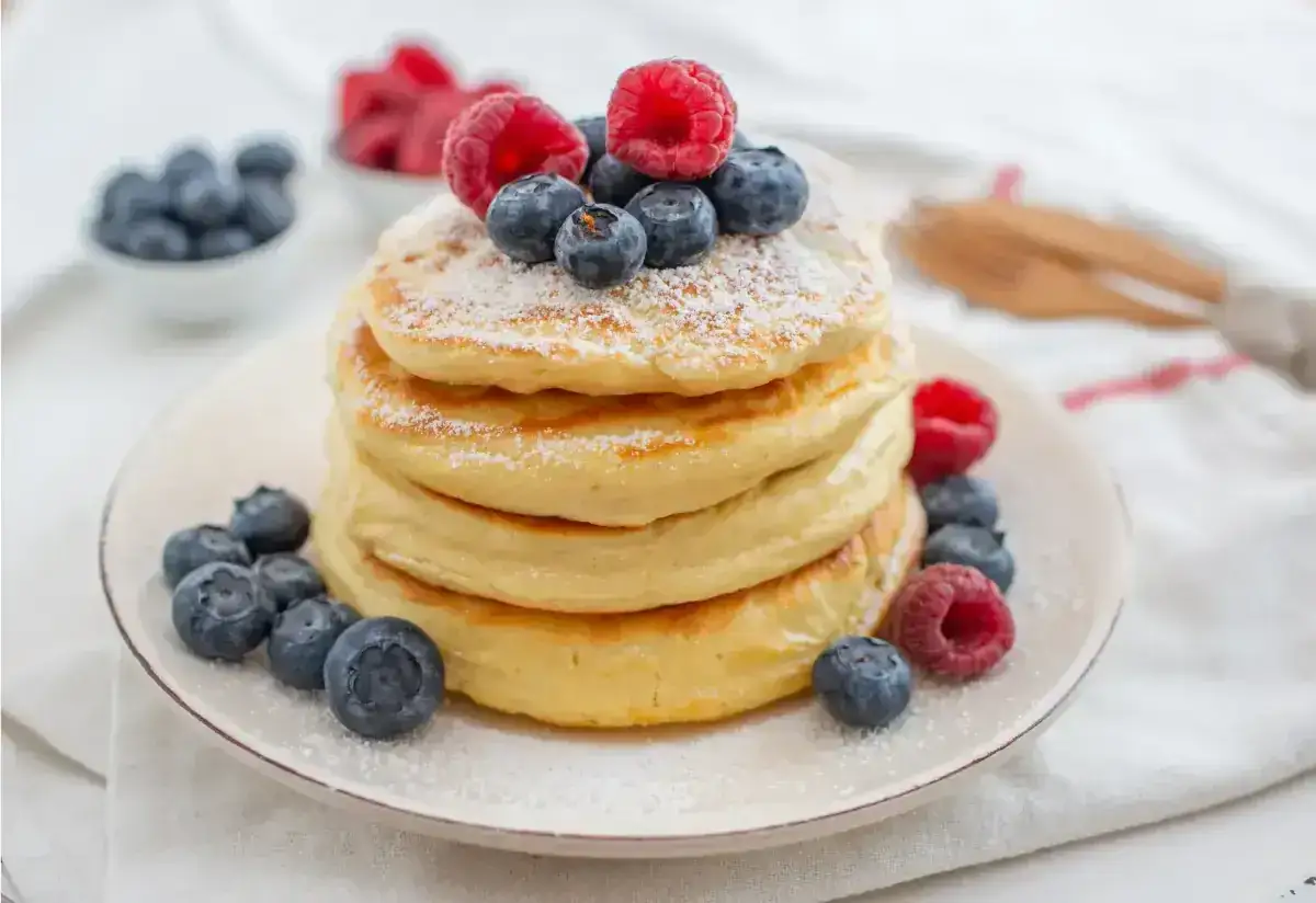 Pile de pancakes sans gluten, garnie de framboises et myrtilles fraîches, saupoudrée de sucre glace.