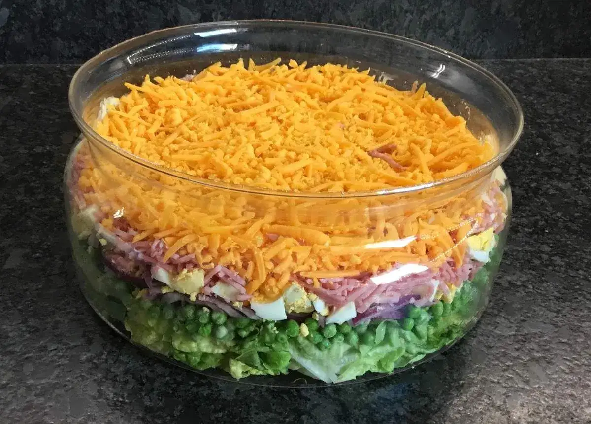 Une salade 7 étages colorée dans un grand bol en verre, avec des couches de laitue, petits pois, oignons rouges, jambon, œufs durs et fromage râpé.