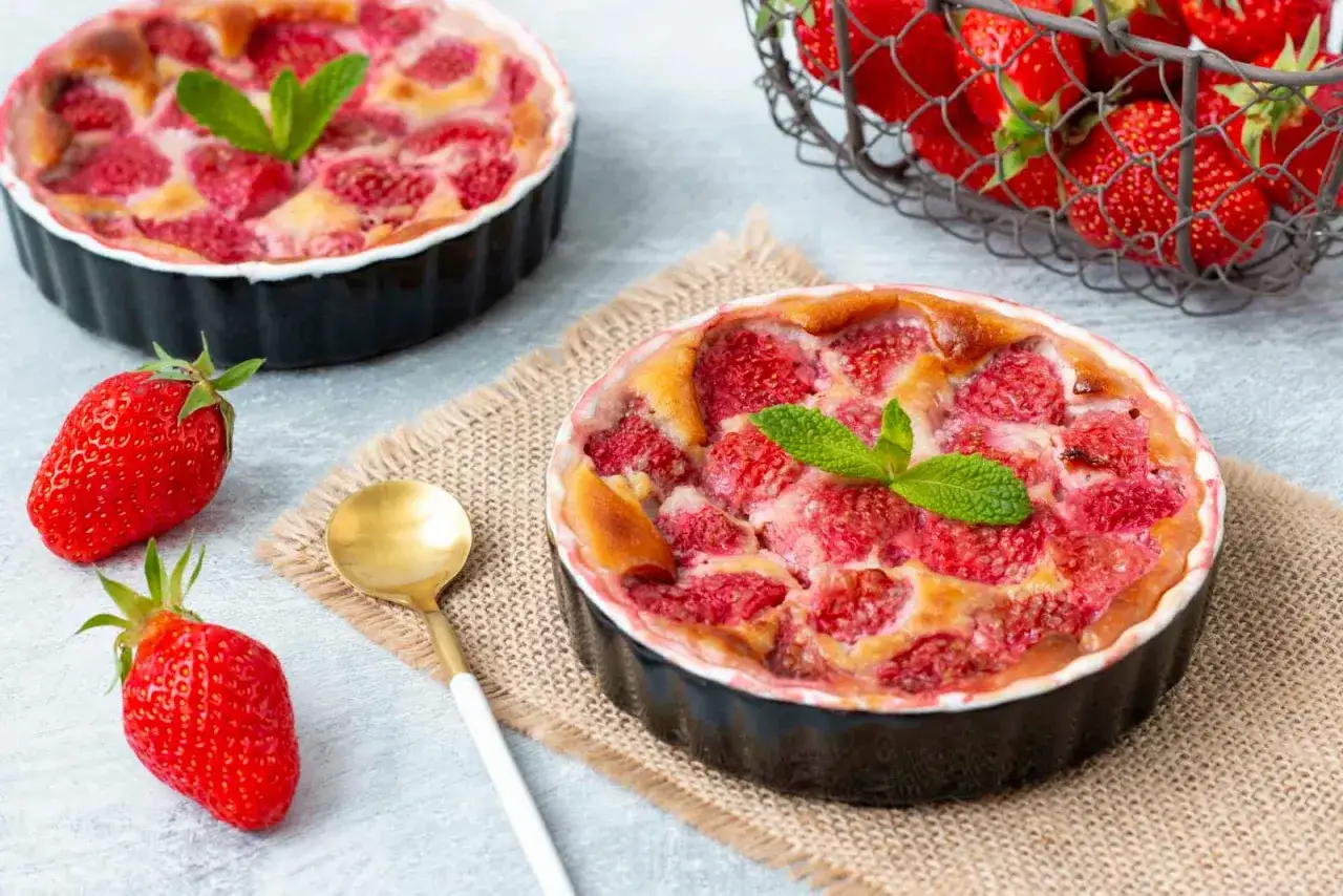 Deux clafoutis aux fraises dorés, ornés de menthe fraîche, prêts à être dégustés. Des fraises fraîches et une cuillère dorée complètent cette scène gourmande.