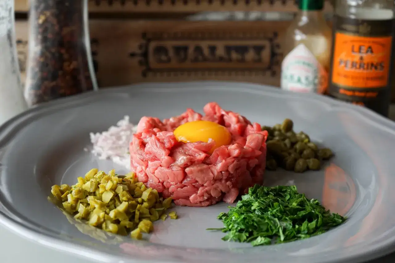 Un plat de tartare de bœuf cru, garni d'un jaune d'œuf, entouré de câpres, cornichons hachés, échalotes et persil.