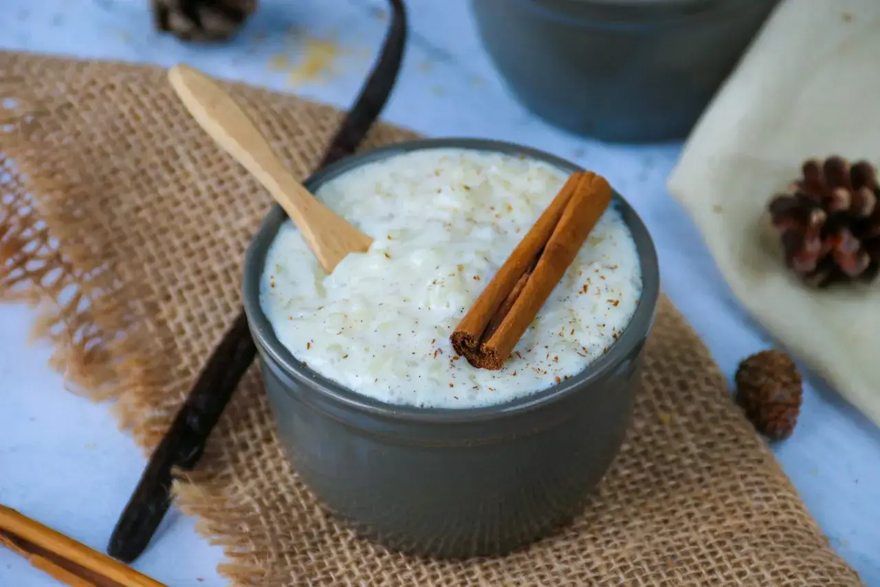 Un bol de riz au lait cr&eacute;meux, garni de b&acirc;tons de cannelle et d'une cuill&egrave;re en bois, sur un fond rustique avec des pommes de pin.