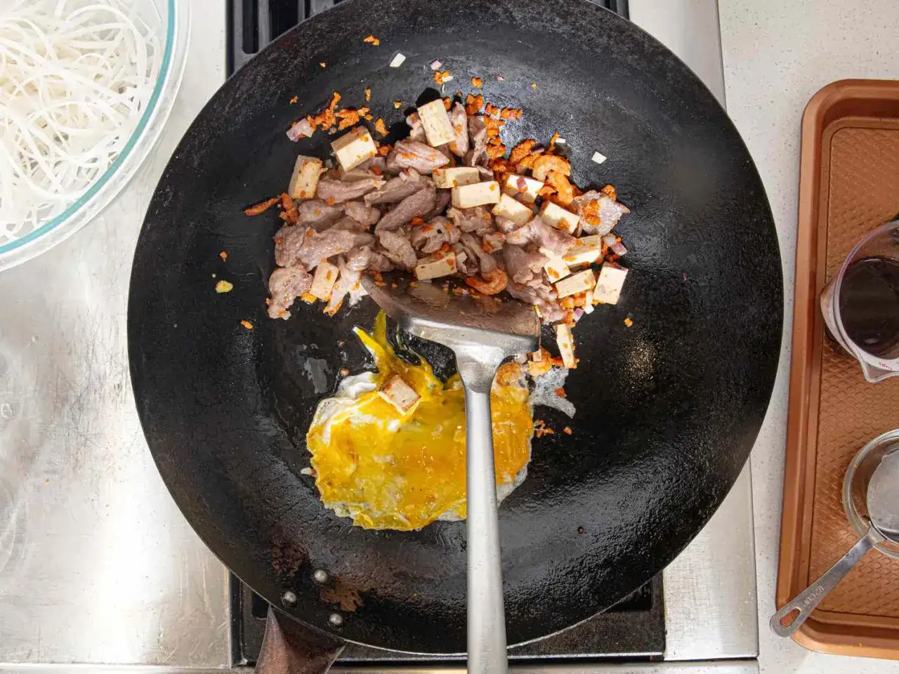 Préparation d'une recette de pad thaï : viande, crevettes, tofu et œuf cuisent dans un wok.