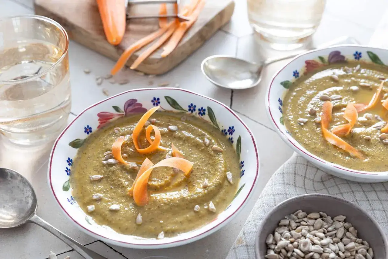 Deux bols de soupe de lentilles, garnie de carottes râpées et de graines de tournesol. Un verre d'eau et des carottes sur une planche complètent la scène.