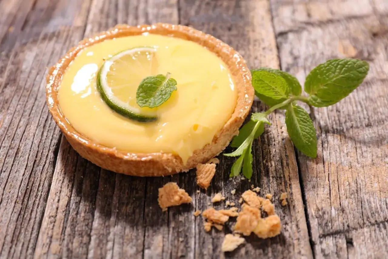 Une tarte aux citrons cr&eacute;meuse, garnie d'une tranche de citron vert et de menthe fra&icirc;che, repose sur un fond de bois rustique.