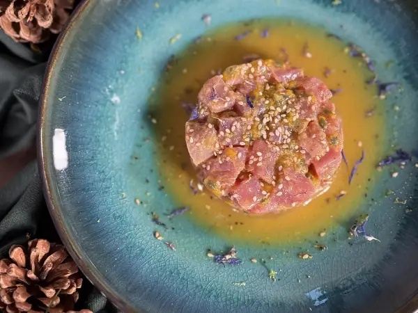 Un délicieux tartare de thon, parsemé de graines de sésame et de fleurs bleues, baignant dans une sauce dorée sur une assiette texturée.